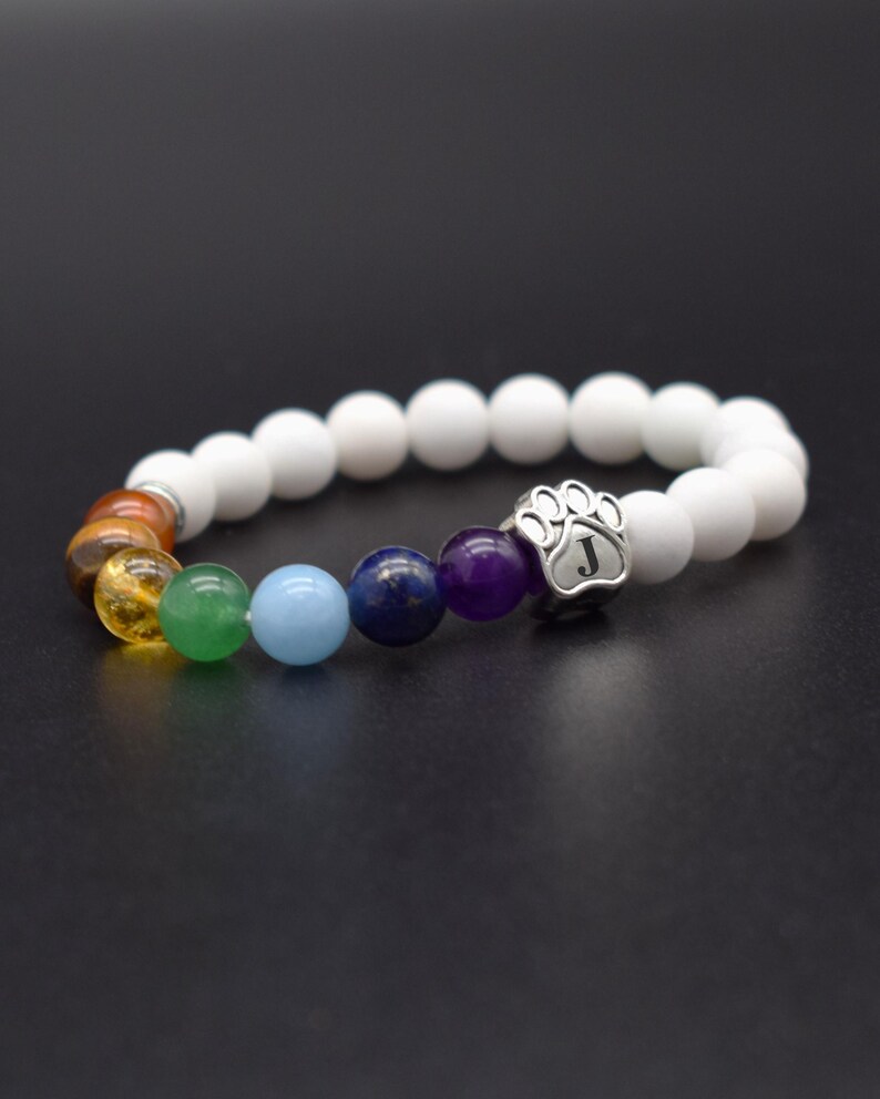 May include: A beaded stretch bracelet featuring matte white beads and a series of colorful gemstone beads in shades of orange, yellow, green, light blue, dark blue, and purple. A silver-toned metal charm shaped like a paw print with the letter J.
