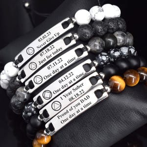 May include: A collection of six beaded bracelets featuring silver-toned metal bars engraved with dates and phrases including Never Give Up, Just for today, One day at a time, 1 Year Sober, and Proud of you DAD, paired with white, black, and tiger eye beads.
