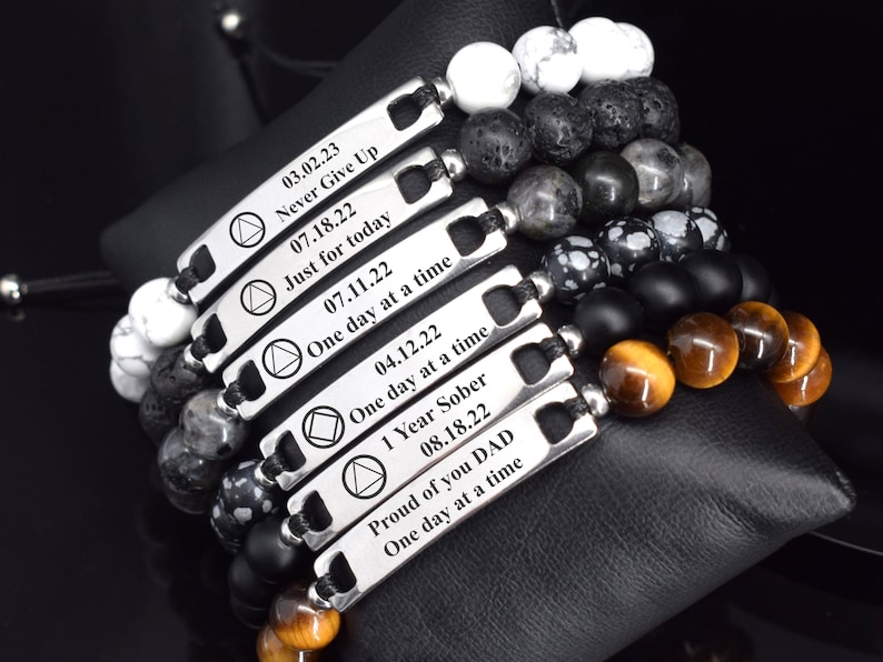 May include: A collection of six beaded bracelets featuring silver-toned metal bars engraved with dates and phrases including Never Give Up, Just for today, One day at a time, 1 Year Sober, and Proud of you DAD, paired with white, black, and tiger eye beads.