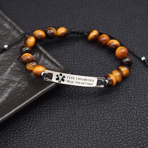 May include: A medical alert bracelet featuring brown and black tiger eye beads on a black cord. A rectangular silver-toned metal plate is engraved with a black medical caduceus symbol, the text TYPE 2 DIABETES, and Mom: 364-647-9647.