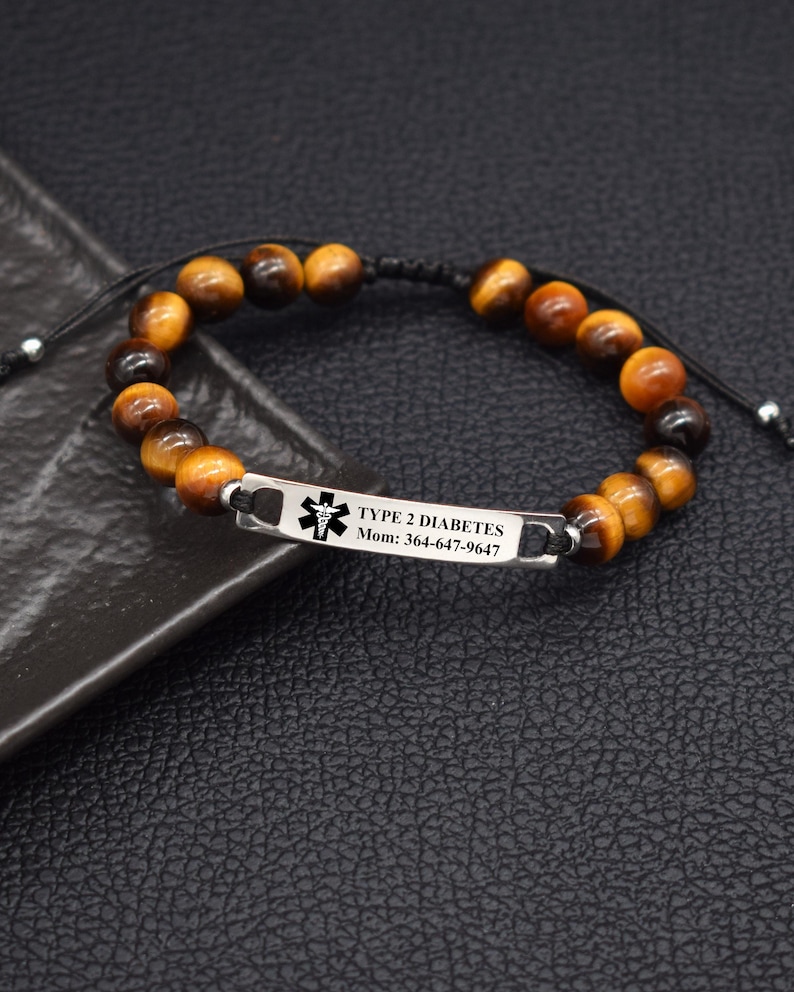 May include: A medical alert bracelet featuring brown and black tiger eye beads on a black cord. A rectangular silver-toned metal plate is engraved with a black medical caduceus symbol, the text TYPE 2 DIABETES, and Mom: 364-647-9647.