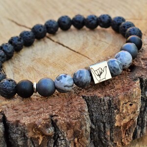 May include: Black lava rock and grey agate beaded bracelet with a silver charm featuring a hand gesture.