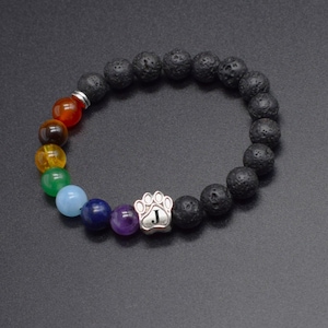 May include: A beaded bracelet featuring black porous lava stone beads and a series of smooth, multi-colored round beads in red, brown, yellow, green, light blue, dark blue, and purple. A silver-toned paw print charm with the letter J is included.