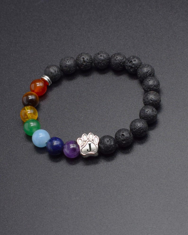 May include: A beaded bracelet featuring black porous lava stone beads and a series of smooth, multi-colored round beads in red, brown, yellow, green, light blue, dark blue, and purple. A silver-toned paw print charm with the letter J is included.