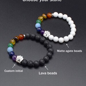 May include: Two beaded bracelets with the text Choose your stone. One features white matte agate beads, the other black lava beads. Both include a sequence of colored chakra beads and a silver paw print charm with a custom initial A or B.