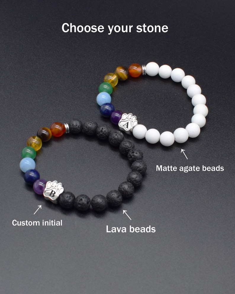 May include: Two beaded bracelets with the text Choose your stone. One features white matte agate beads, the other black lava beads. Both include a sequence of colored chakra beads and a silver paw print charm with a custom initial A or B.