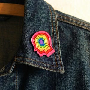 Puede incluir: Un colorido pin de esmalte con forma de cabeza humana, con un diseño en capas con colores del arcoíris. El pin está sujeto a una chaqueta vaquera, creando un contraste vibrante. El diseño del pin es un perfil de cabeza con capas concéntricas.