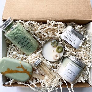 May include: A gift box filled with green and gold themed bath and body products. The box contains a matcha shower creamer, a matcha body bar, a matcha lotion shake, a candle with green sprinkles, and a wooden stick. The products are all packaged in small, silver tins with white labels.