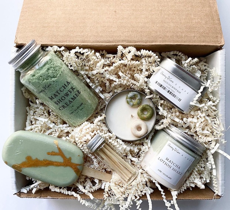 May include: A gift box filled with green and gold themed bath and body products. The box contains a matcha shower creamer, a matcha body bar, a matcha lotion shake, a candle with green sprinkles, and a wooden stick. The products are all packaged in small, silver tins with white labels.