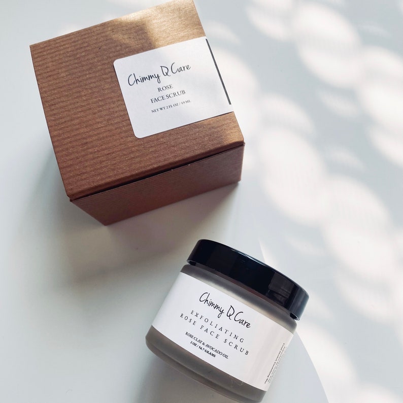 K&ouml;nnte beinhalten: Ein brauner Karton mit einem wei&szlig;en Etikett, auf dem "Chimmy Q Care Rose Face Scrub Net Wt 1 fl oz / 30 ml" steht. Ein Glasgef&auml;&szlig; mit schwarzem Deckel und einem grauen Etikett, auf dem "Chimmy Q Care Exfoliating Rose Face Scrub Rose Clay & Avocado Oil 1.0 fl oz / 30 ml" steht, befindet sich auf einer wei&szlig;en Oberfl&auml;che.