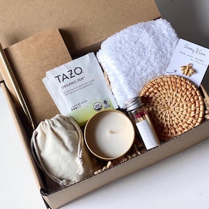 May include: A gift box filled with various items, including a small notebook, a tea bag, a candle, a small bag, a white towel, a small glass jar with matches, and a woven coaster. The box is made of cardboard and has a brown color.