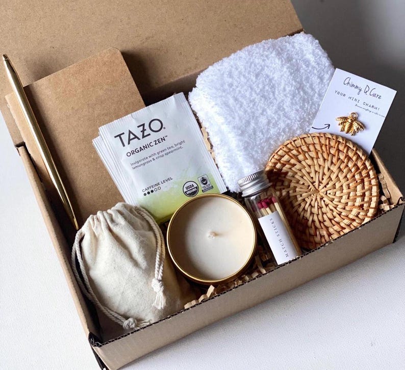 May include: A gift box filled with various items, including a small notebook, a tea bag, a candle, a small bag, a white towel, a small glass jar with matches, and a woven coaster. The box is made of cardboard and has a brown color.