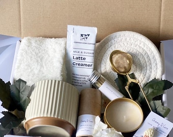 May include: A white gift box filled with various items, including a white towel, a small white ceramic pot, a brown cardboard tube, a gold candle, a gold tea strainer, a white coaster, a small glass bottle, a white and gold package of coffee creamer, and a small white card with the text "Chimmy Cure" and a picture of an ice cream cone.