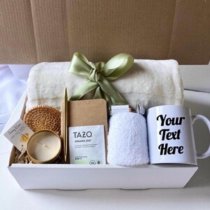 May include: A white gift box filled with a variety of items, including a white throw blanket, a white mug with the text "Your Text Here", a candle, a small notebook, a pen, a tea bag, a washcloth, and a woven coaster.