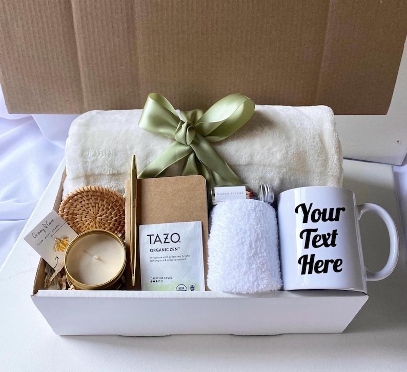May include: A white gift box filled with a variety of items, including a white throw blanket, a white mug with the text "Your Text Here", a candle, a small notebook, a pen, a tea bag, a washcloth, and a woven coaster.
