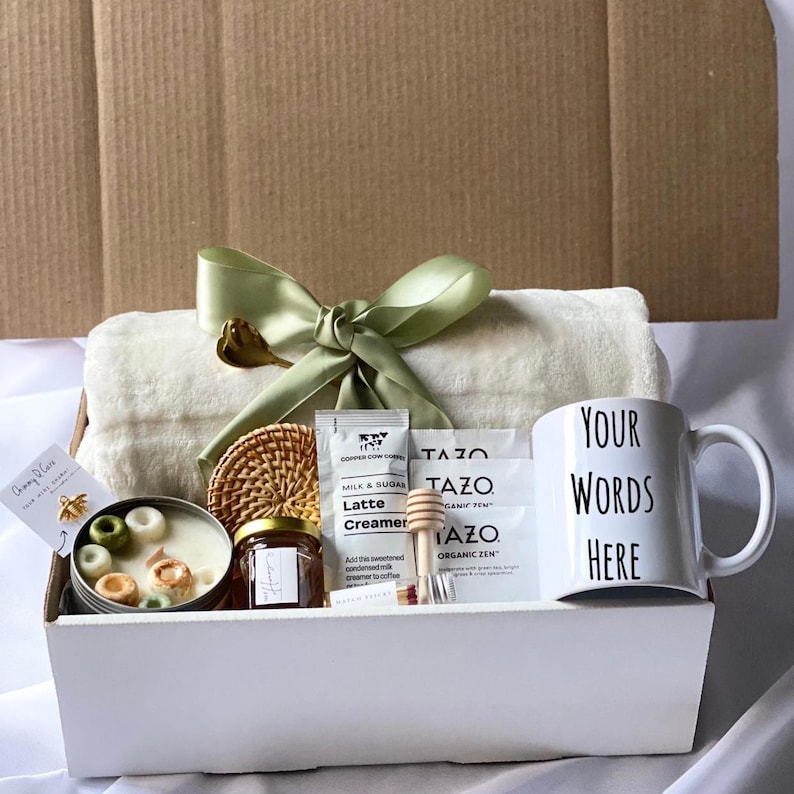 May include: A white gift box filled with a variety of items, including a white blanket, a mug with the text "YOUR WORDS HERE", a jar of honey, a candle, a package of coffee creamer, and two tea bags. The box is tied with a green ribbon.