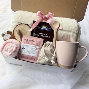 May include: A gift box filled with self-care items. Includes a pink mug, a soft blanket, hot cocoa mix, tea, honey, a wooden spoon, and a pink face towel. The box is tied with a pink ribbon.