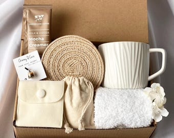 Puede incluir: Una caja de regalo marrón llena de una taza blanca, un posavasos tejido marrón, una toalla blanca, una pequeña bolsa blanca, un pequeño sobre blanco con un botón y un pequeño paquete marrón con el texto "Copper Cow Coffee Milk & Cocoa Mocha Creamer" en él.