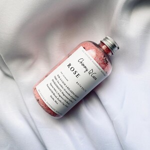 Może przedstawiać: Szklany słoik wypełniony r&oacute;żowymi solami do kąpieli z etykietą "Chimmy Care Rose Bath Salts" i srebrną pokrywką. Słoik znajduje się na białym tle z tkaniny. Etykieta zawiera r&oacute;wnież tekst "All Natural Handmade Skincare" i "For a Calm a Relaxing Bath".
