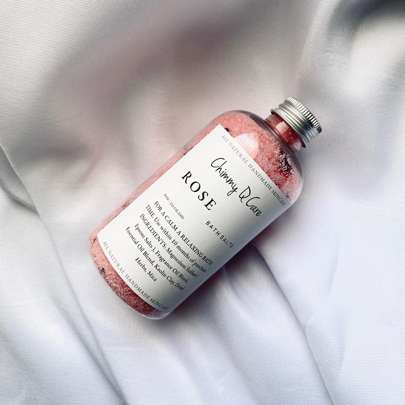Może przedstawiać: Szklany słoik wypełniony r&oacute;żowymi solami do kąpieli z etykietą "Chimmy Care Rose Bath Salts" i srebrną pokrywką. Słoik znajduje się na białym tle z tkaniny. Etykieta zawiera r&oacute;wnież tekst "All Natural Handmade Skincare" i "For a Calm a Relaxing Bath".