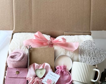 May include: A white gift box filled with a variety of items, including a pink blanket, a pink headband, a pink candle, a white mug, a pink pouch, a pink and white pin, and a small bag of pink fabric. The box is decorated with a pink ribbon.