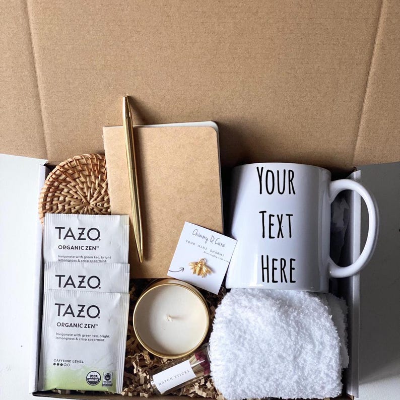 May include: A gift box filled with items including a white mug with the text "YOUR TEXT HERE", a small candle, a notebook, a pen, two tea bags, a coaster, a small white towel, and a gold bee charm.