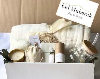 May include: A white gift box filled with a variety of items, including a white blanket, a wooden coaster, a candle, a small bag, a glass jar with a straw, and a small tube of matches. The box is decorated with a white ribbon and a card that reads "Eid Mubarak, Taqabal Allah minna wa minkum".