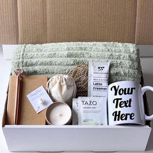 May include: A white gift box filled with a green towel, a candle, a small bag, a notebook with a pen, a coaster, a coffee creamer packet, a tea bag, and a white mug with the text "Your Text Here".