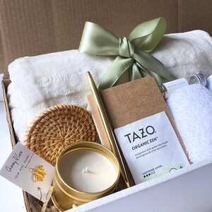 May include: A white gift box filled with a white towel, a gold pen, a brown notebook, a wicker coaster, a gold candle, a tea bag, a white washcloth, and a small glass bottle with matches. The box is tied with a green satin ribbon.