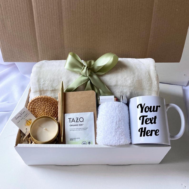May include: A white gift box filled with a white throw blanket, a mug that says "Your Text Here", a candle, a small notebook, a tea bag, a small jar of matches, a coaster, and a white washcloth.