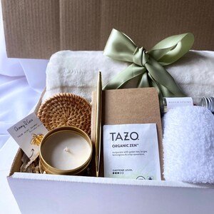 May include: A white gift box filled with a variety of items, including a white towel, a gold pen, a brown notebook, a gold candle, a woven coaster, a tea bag, and a small bottle of match sticks. The box is tied with a green satin ribbon.
