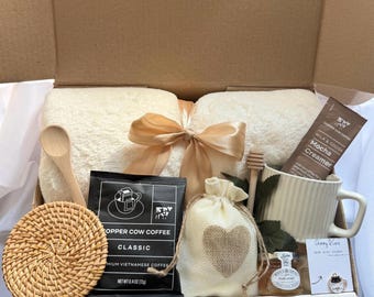 Puede incluir: Una caja de regalo con una manta color crema atada con una cinta dorada, una taza, café, miel y un posavasos. La caja incluye una bolsa de Copper Cow Coffee, un sobre de crema de moca y una pequeña bolsa con un corazón.