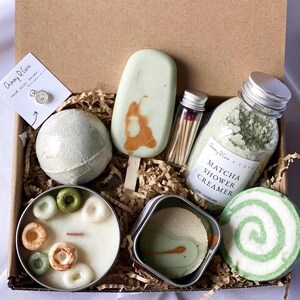 May include: A green and white gift box filled with bath and body products. The box includes a green bath bomb, a green and white swirl bath bomb, a green shower cream, a green soap bar, a green candle, a small tin of green bath salts, and a small jar of matches. The box is labeled "Chimmy D'Care Your Mini Spa Gift".