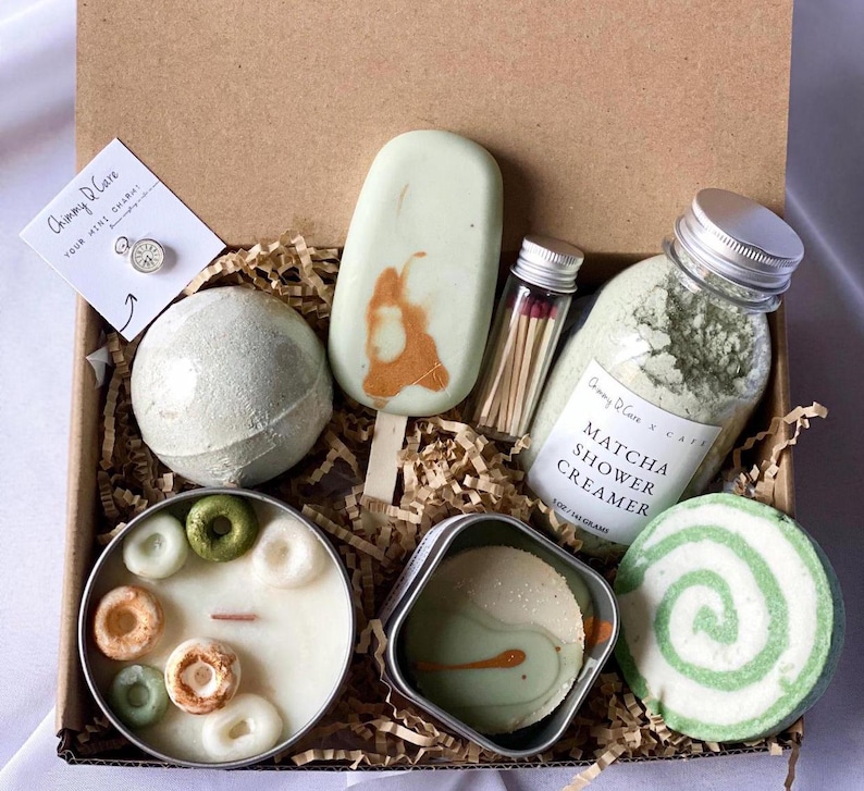 May include: A green and white gift box filled with bath and body products. The box includes a green bath bomb, a green and white swirl bath bomb, a green shower cream, a green soap bar, a green candle, a small tin of green bath salts, and a small jar of matches. The box is labeled "Chimmy D'Care Your Mini Spa Gift".
