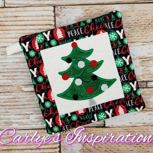 May include: A black and white quilted pot holder with a green Christmas tree design in the center. The border of the pot holder has a repeating pattern of red and white snowflakes, Christmas lights, and the words "Peace", "Joy", "Merry", and "Cheer".