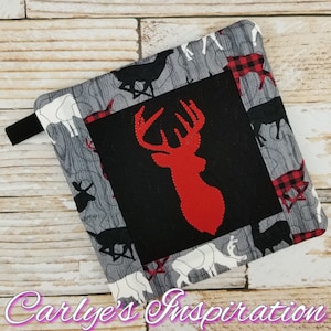 Puede incluir: Un agarrador de olla acolchado rojo y negro con una silueta de ciervo en el centro. El agarrador de olla está rodeado por un borde de tela a cuadros blanco y rojo y una tela gris con un patrón de veta de madera.