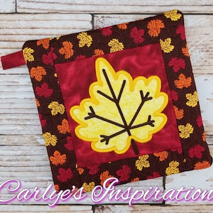 May include: A red quilted pot holder with a yellow embroidered leaf in the center. The pot holder is surrounded by a brown fabric with a pattern of small yellow and orange leaves.