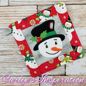 May include: Red, white, and green quilted pot holder with a snowman face in the center. The snowman has a black top hat, green holly, and a carrot nose. The pot holder is surrounded by smaller snowmen.