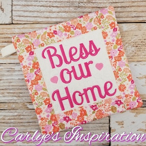 May include: A square, quilted pot holder with a floral pattern in pink, orange, and green. The center features the embroidered words "Bless our Home" in pink, with two pink hearts. A small loop is attached to the side.