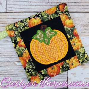 May include: A black and orange pot holder with a pumpkin design. The pumpkin is embroidered with a detailed design and has a green stem. The pot holder is surrounded by a border of green leaves and pumpkins.