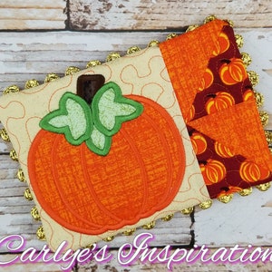 May include: A small, quilted cushion with a gold trim features an embroidered orange pumpkin with green leaves. The background fabric is cream with a swirl pattern. The cushion is on a wooden background.