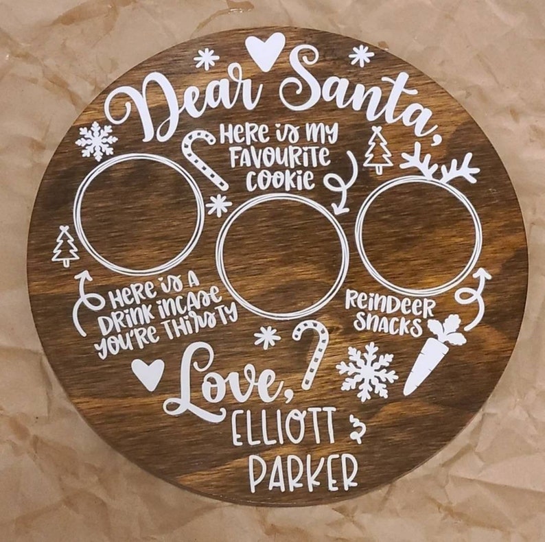 May include: A brown wooden round plate with white text and images. The text reads "Dear Santa, Here is my favourite cookie, Here is a drink incase you're thirsty, Love, ELLIOTT PARKER". The plate has three circles for placing treats and images of a candy cane, a snowflake, a carrot, a Christmas tree, and a star.