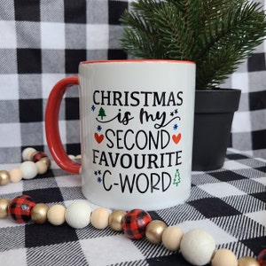 Peut inclure: Mug en céramique blanche avec une anse et un bord intérieur rouges. Le mug affiche le texte "CHRISTMAS is my SECOND FAVOURITE *C-WORD" en noir, avec des accents festifs. Un petit conifère en pot est en arrière-plan.