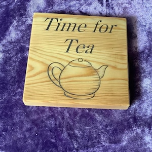May include: A square wooden sign with the words "Time for Tea" in black lettering. A black outline of a teapot is below the text. The sign is made of light-coloured wood with visible grain.