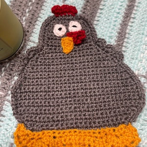 Peut inclure: Un poulet en crochet gris avec des yeux rouges et blancs, un bec jaune et une crête rouge. Le poulet a une base en crochet jaune.