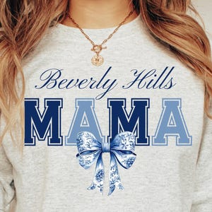 以下が含まれることがあります： グレーのスウェットシャツに、青い文字で「Beverly Hills Mama」と書いてあります。テキストの下には、花柄の青いリボンが中央に配置されています。