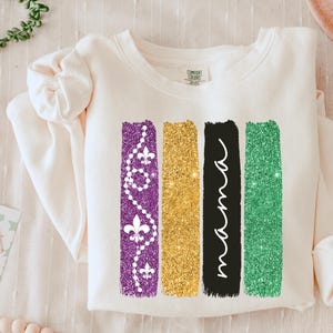 Puede incluir: Una sudadera blanca con un diseño de tres rayas verticales en morado, dorado y verde. Las rayas son brillantes y tienen un efecto de pinceladas. La raya morada tiene un diseño de flores de lis y cuentas. La raya dorada es sólida. La raya verde tiene un diseño de líneas onduladas.