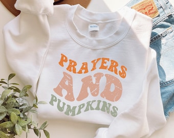 Puede incluir: Sudadera blanca con el texto "Prayers and Pumpkins" en naranja, melocotón y verde. La sudadera está doblada con unos pantalones cortos vaqueros y un sombrero marrón en el fondo.