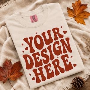Può includere: Una t-shirt a maniche corte color crema con il testo YOUR DESIGN HERE in un carattere arancione scuro, audace e ondulato. Il design è accentuato da piccole stelle e cuori arancioni scuri. L'etichetta Comfort Colors è presente sul collo.