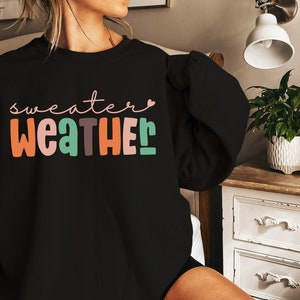 以下が含まれることがあります： カラフルで遊び心のある文字で「sweater weather」と書かれた黒いスウェットシャツ。テキストにはオレンジ、ピンク、グリーン、ブラウンの色が含まれています。「sweater」という単語は筆記体で書かれており、「r」の上には小さなピンク色のハートがあります。
